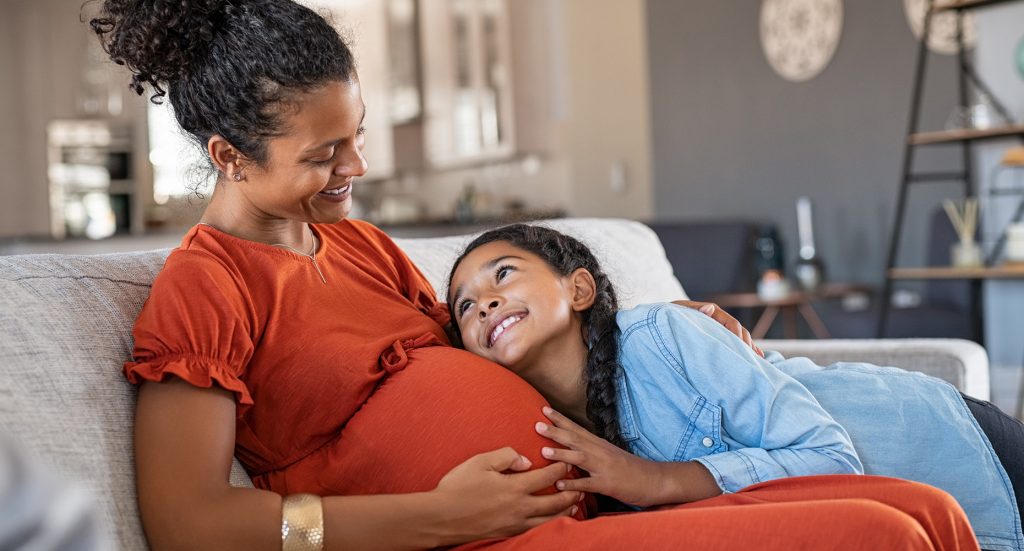 pregnant mother and daughter smiling at each other sitting on couch